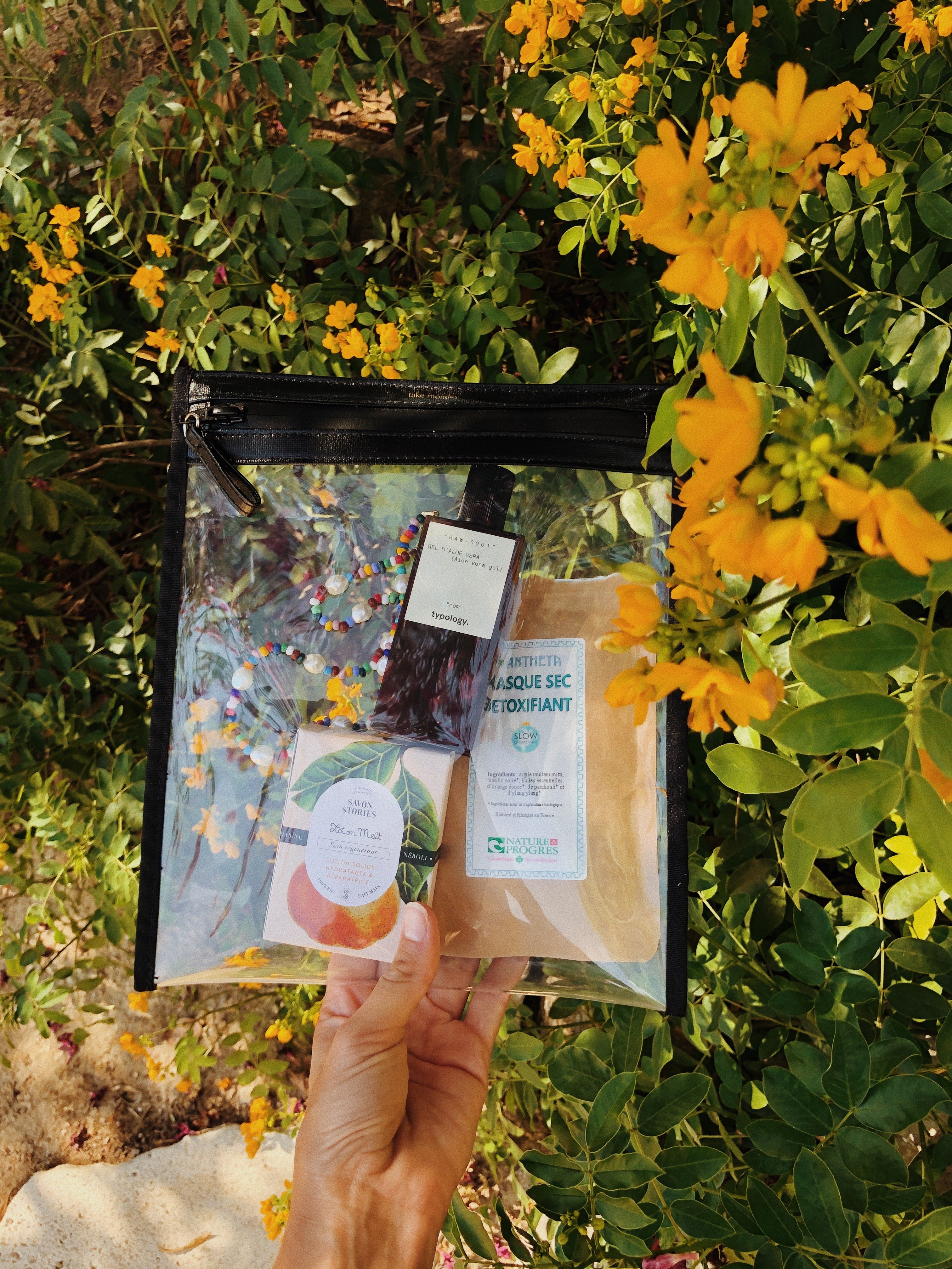 Photograph of a hand holding a transparent beauty pouch containing Typology Aloe Vera Gel, a Savon Stories peach-labeled lotion bar, and a colorful beaded pearl bracelet, set against a lush background of vibrant yellow flowers and green foliage