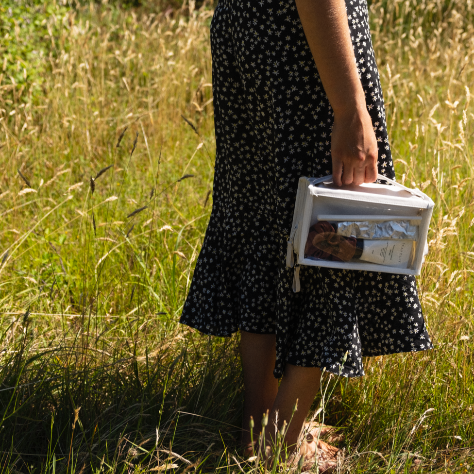 A person wearing a black dress with white floral patterns stands in a sunlit golden grassy field holding a clear white cosmetic bag revealing a brown scrunchie a silver foil packet and a cream tube with SEA OLLY visible text