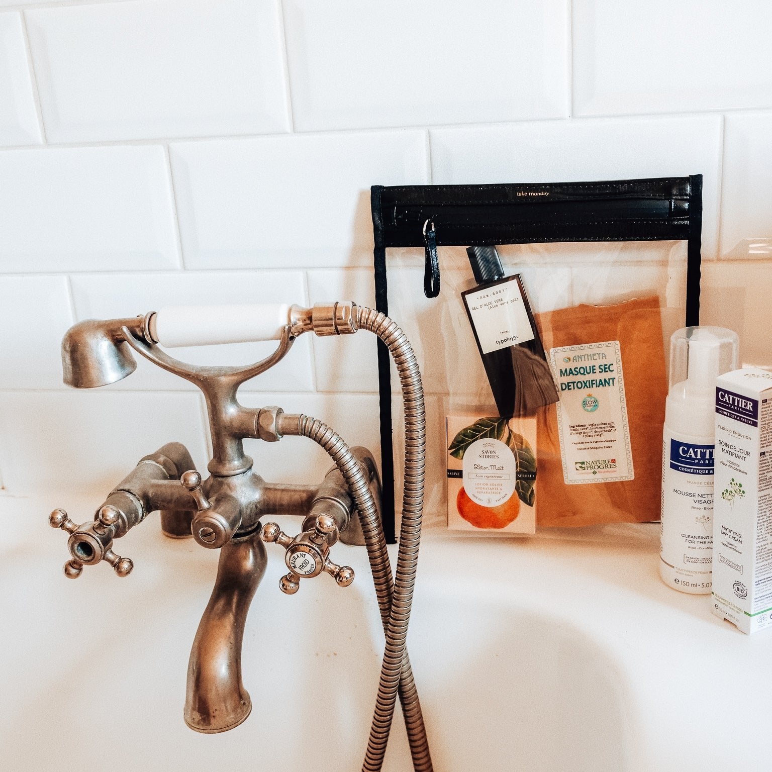 A white-tiled bathroom scene showcasing a vintage bronze bathtub faucet, a clear 'take monday' toiletry bag holding Typology and Antheta products, alongside Cattier and Savon Stories skincare with an orange label