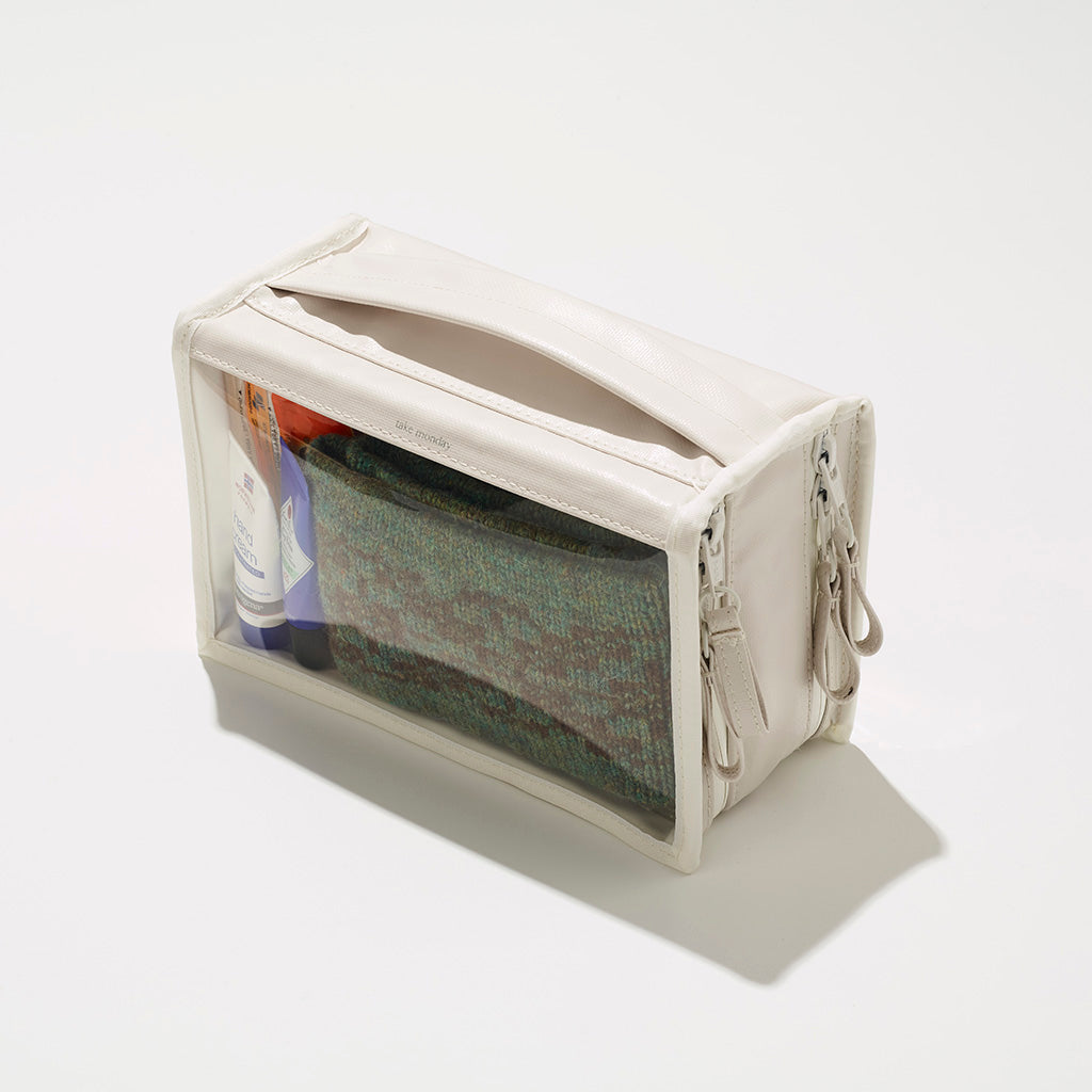 Off-white take monday clear front travel bag containing a folded green speckled garment and a hand cream bottle on a white studio background