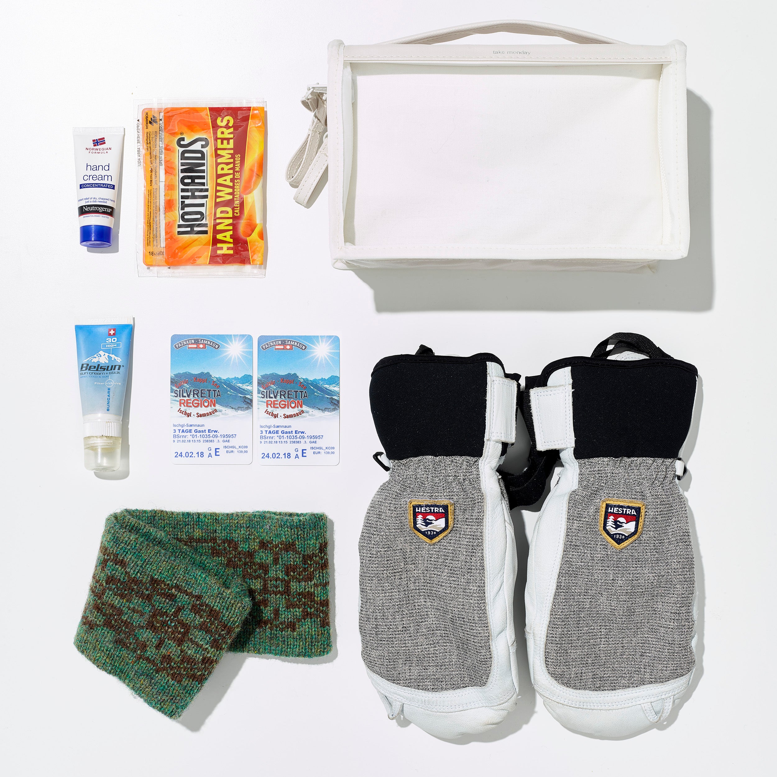 Flat lay photo of winter skiing essentials for the Silvretta Ischgl-Samnaun region including white Hestra mittens, bright orange HotHands warmers, a green and brown knit scarf, and skincare products