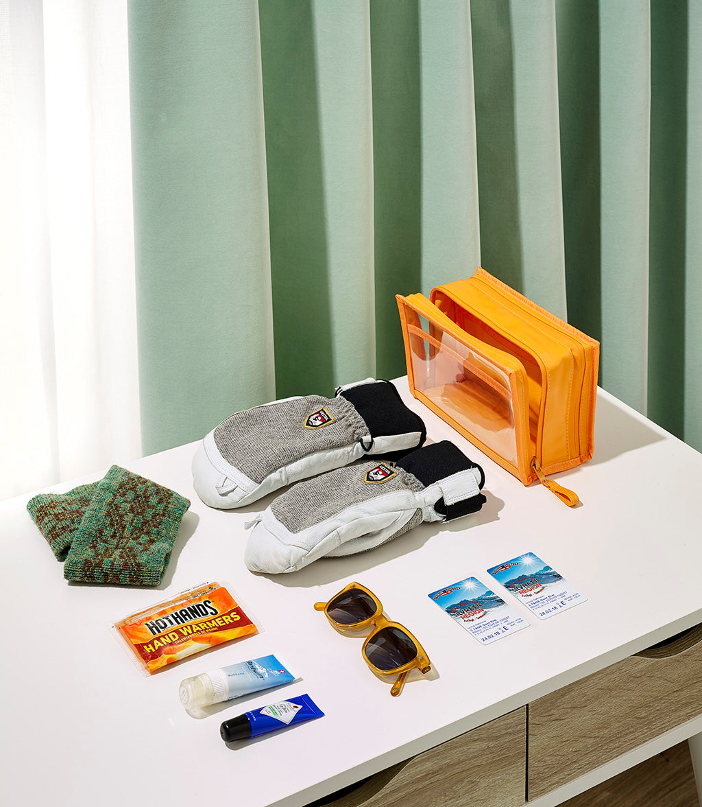 Winter travel essentials arranged on a white table including an orange transparent zipper pouch, gray and white ski mittens with a resort logo, green knit fingerless gloves, yellow sunglasses, a Hothands hand warmers packet, and two Silvretta Montafon ski passes