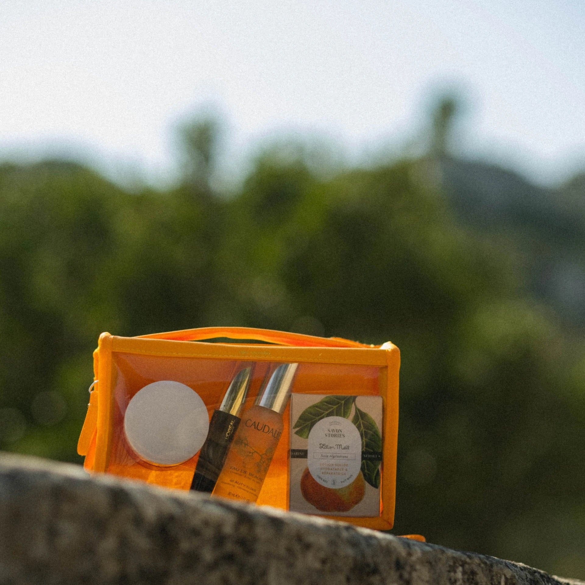 Photography of an orange transparent travel cosmetics bag filled with Caudalie Eau de Beauté, a Savon Stories pear-graphic soap bar, and other skincare items, resting on a stone ledge against a blurred green hilly landscape
