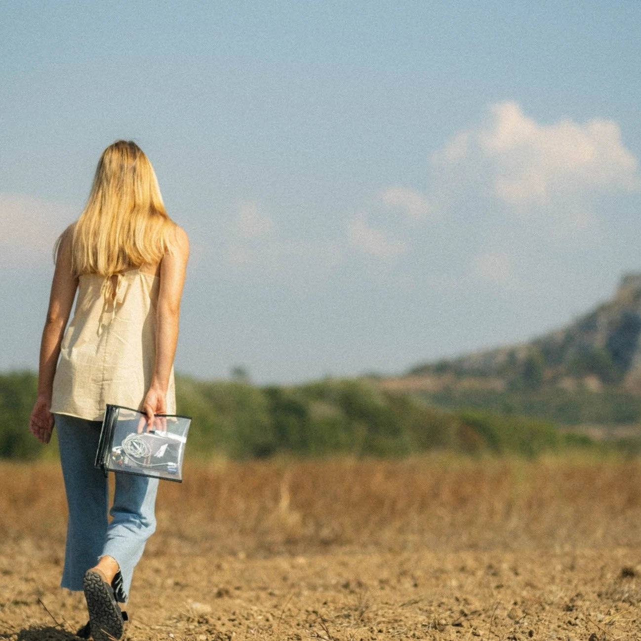 Photography of a blonde woman with long blonde hair in a beige backless top and light blue jeans walking away across dry cracked earth, holding a clear transparent pouch with electronic cables, towards distant green trees and brown hills under a pale blue sky