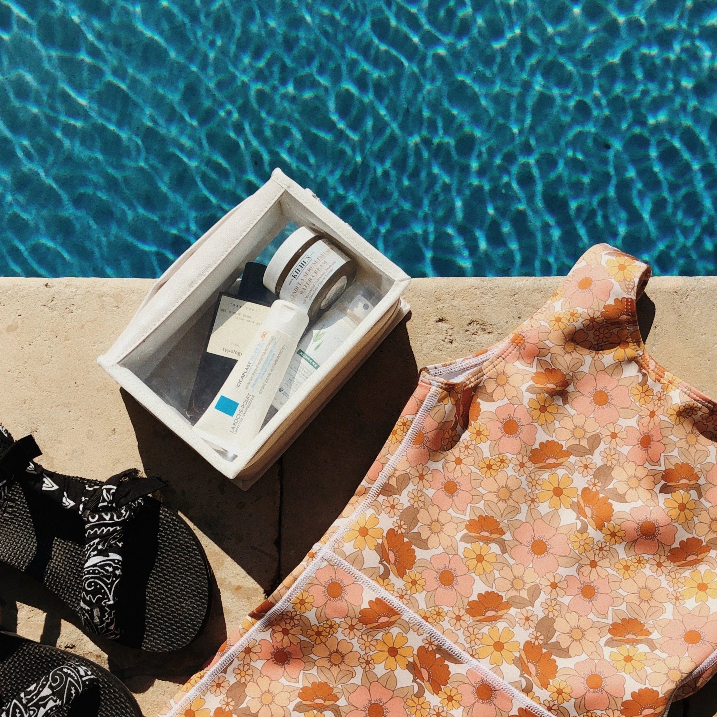 Photography of poolside essentials featuring a retro floral swimsuit black Teva sandals and a clear bag of Kiehl's skincare beside sparkling blue water