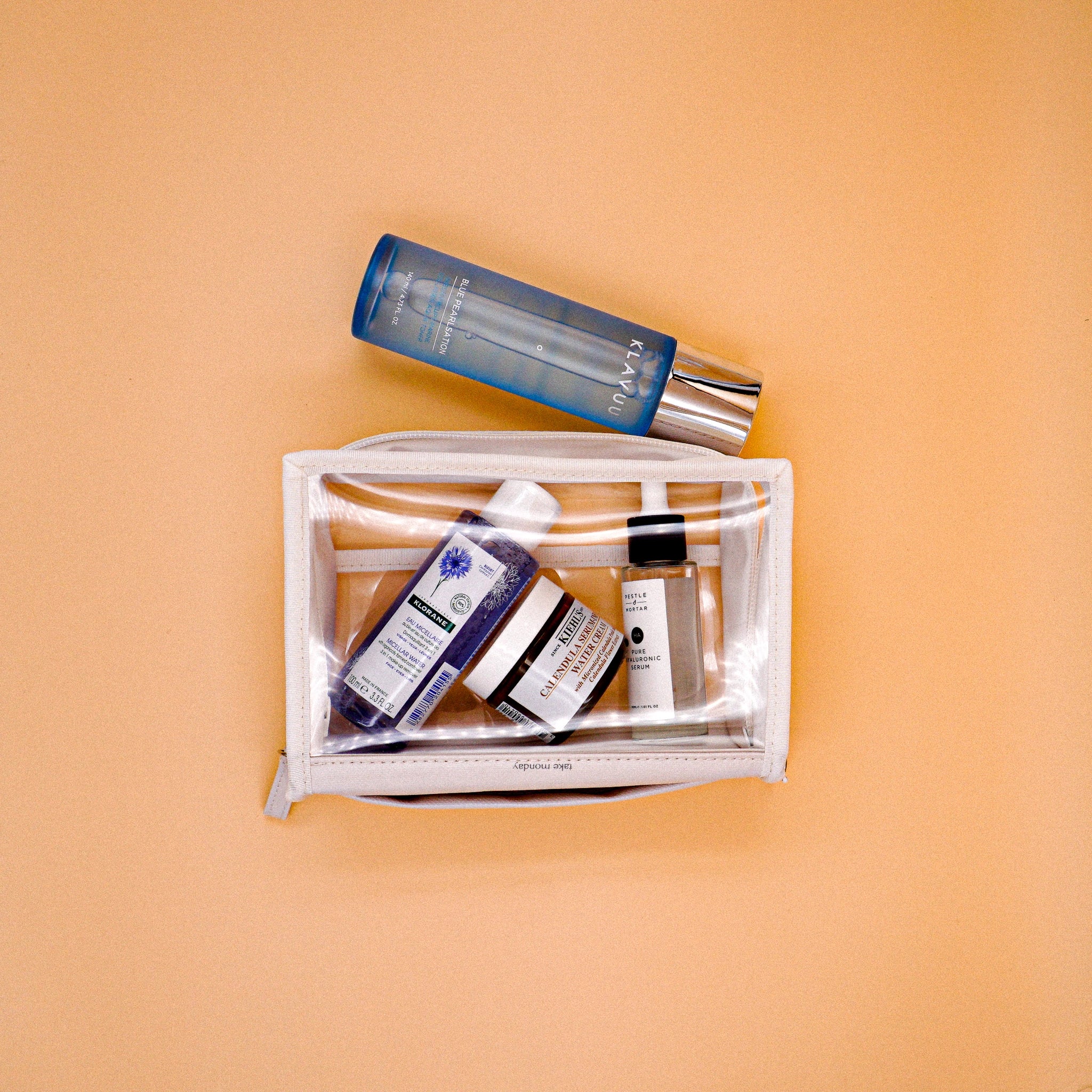 Flat lay of various skincare products including a blue Klavuu toner and a clear cosmetic pouch holding Kiehl's Calendula Cream, Klorane Micellar Water, and Pestle & Mortar Hyaluronic Serum on a peach background