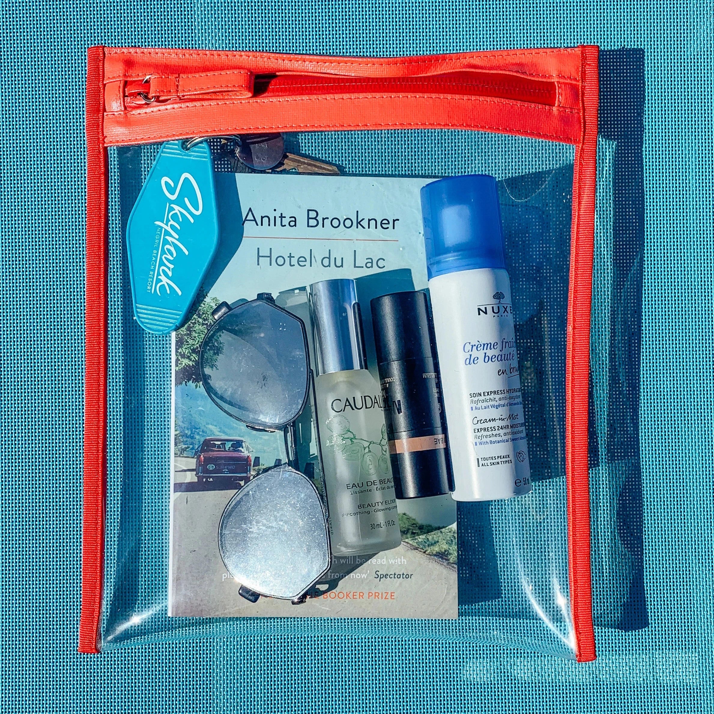 Photograph of a clear travel pouch with vibrant orange trim, containing a bright blue Skylark Negril Beach Resort keychain, Anita Brookner's Hotel du Lac book, and reflective sunglasses on a textured blue mat