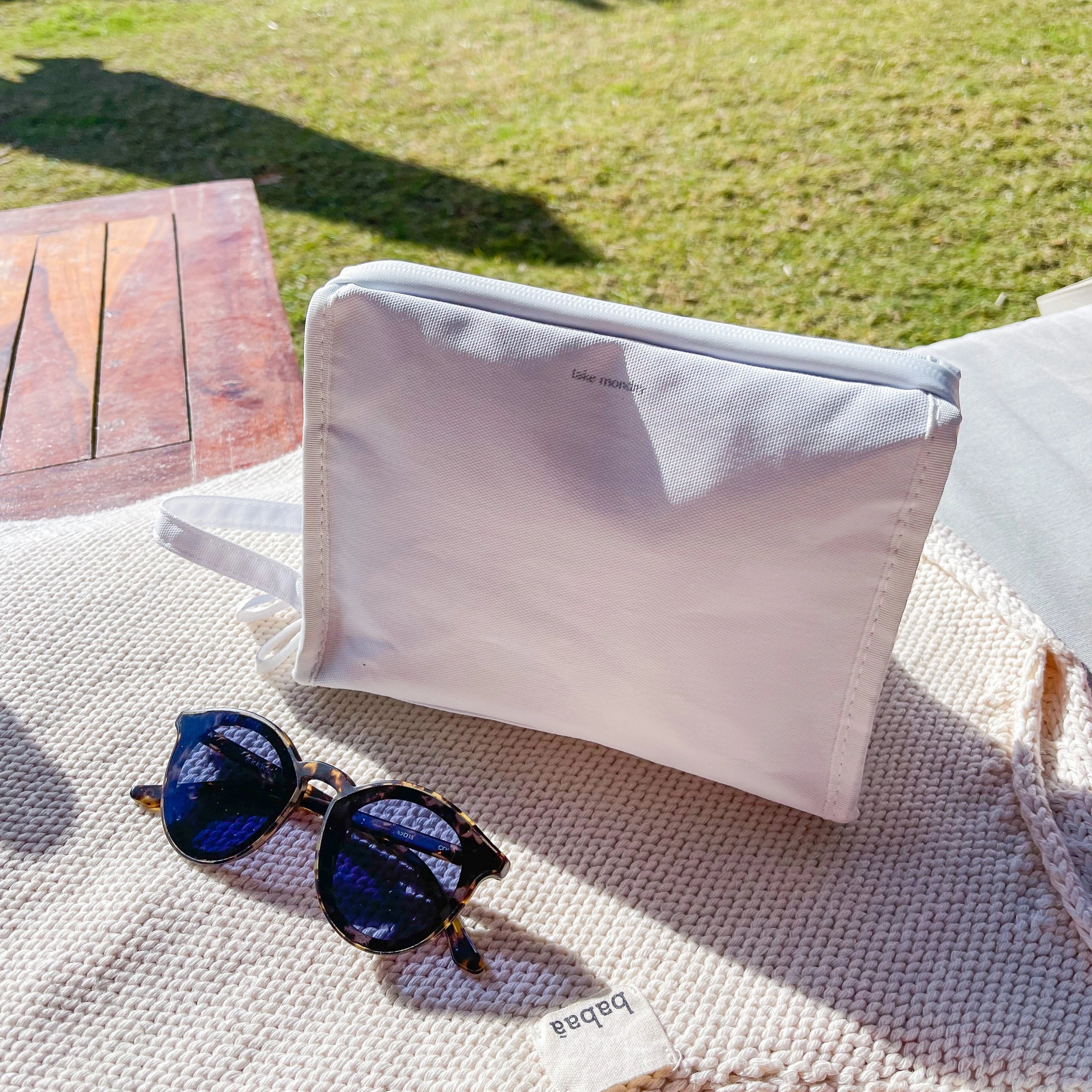 A white take monday brand pouch and tortoiseshell sunglasses with dark lenses rest on a cream babaà knitted mat outdoors in bright sunlight with green grass and a wooden table