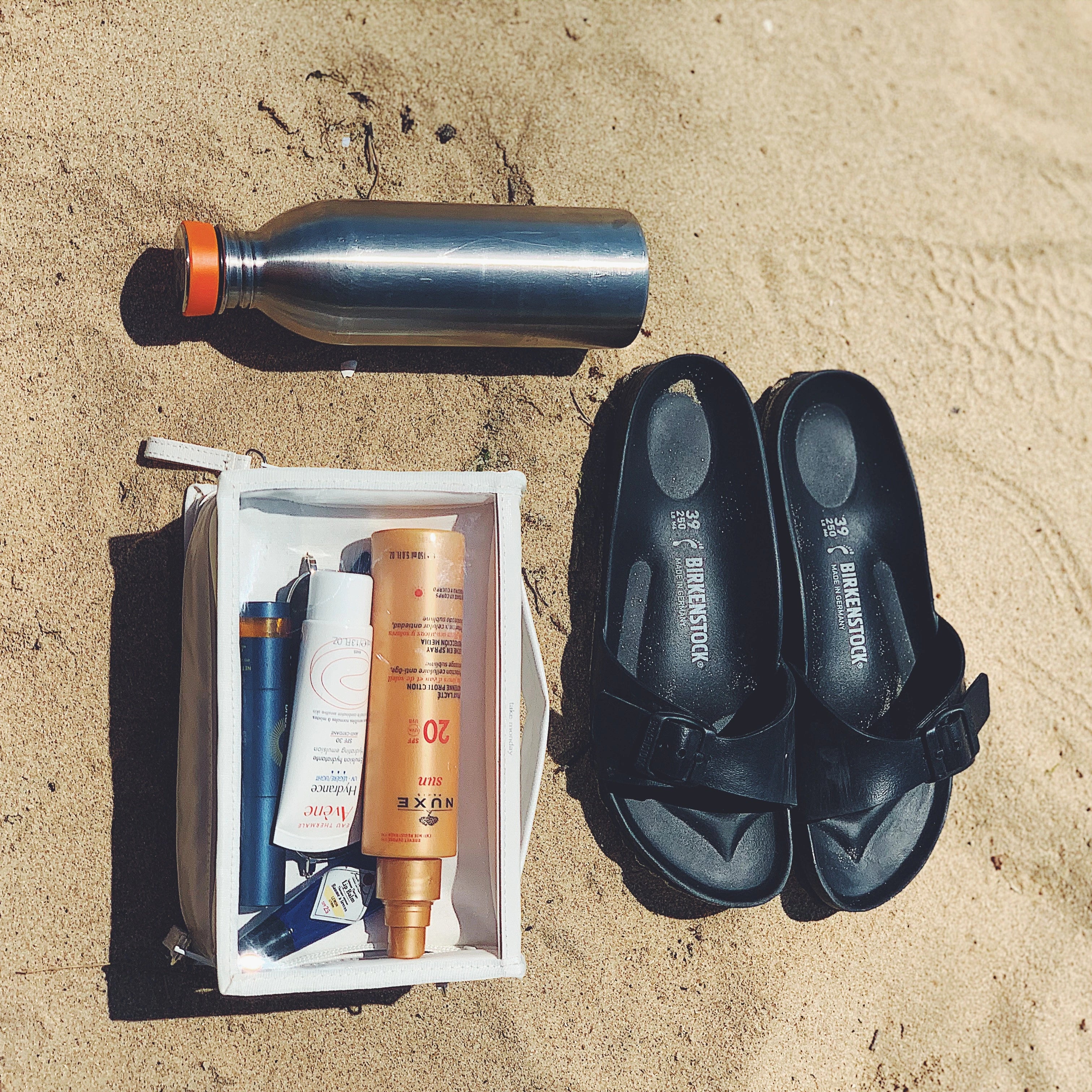Flat lay photograph of beach essentials on golden sand featuring a silver water bottle with an orange cap, black Birkenstock sandals, and a white toiletry bag displaying Nuxe SPF 20, Avène SPF 30, and blue lip balm