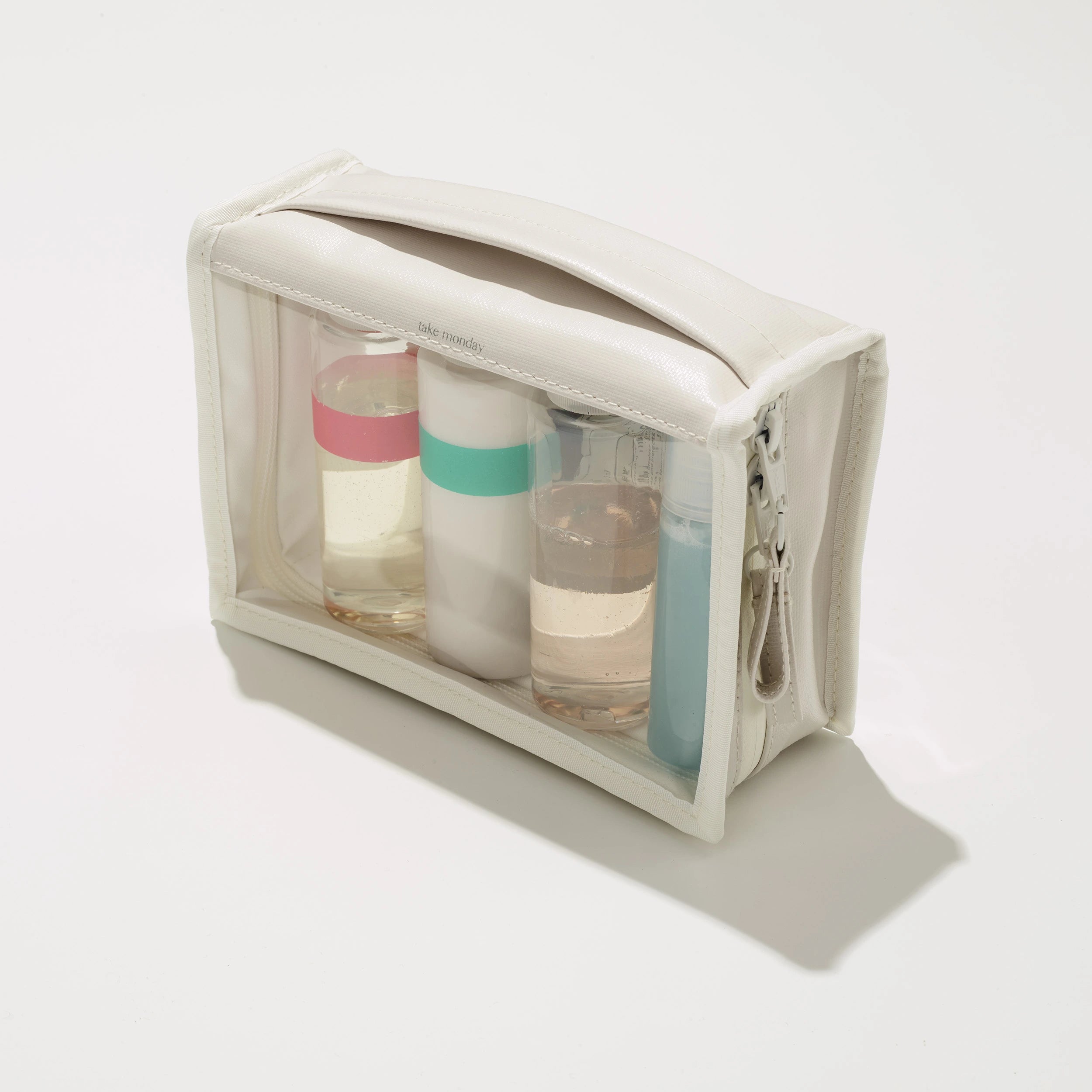 A transparent take monday travel toiletry bag holds four bottles, one with a pink band and another with a teal band, on a bright white studio surface
