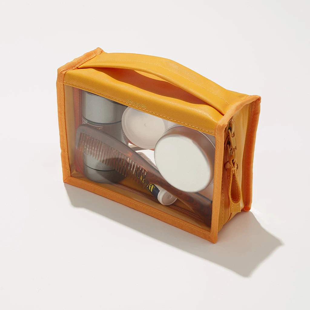 Photograph of a vibrant orange and clear PVC travel toiletry bag containing a wooden comb and various white and silver travel-sized containers on a white studio background