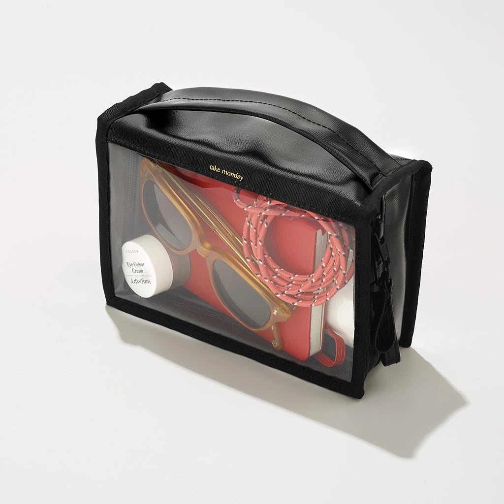 A "take monday" branded clear-front black travel pouch containing amber sunglasses, a vibrant red notebook, and a coiled red and white rope, resting on a clean white background