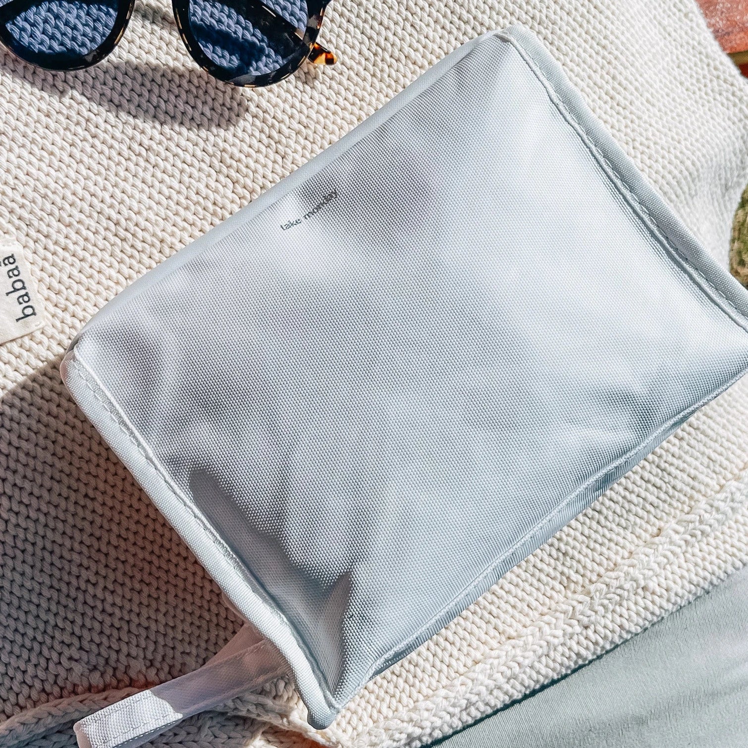 A light blue textured pouch printed with "take monday" rests on a cream chunky knit "babaa" blanket beside tortoiseshell sunglasses with blue reflective lenses under bright sunlight